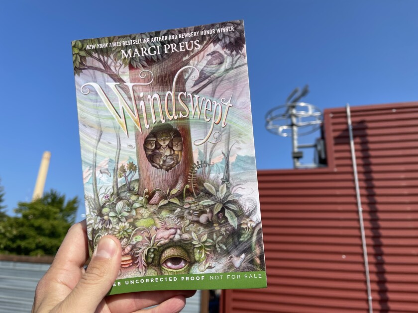Hand holding paperback book: Margi Preus, "Windswept." In background, red building with wind vane is visible out of focus.