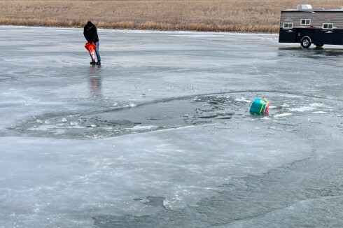 A hole in the ice in on a lake with a small cooler floating and a man placing cones beside the opening. A fishhouse sits in the background.
