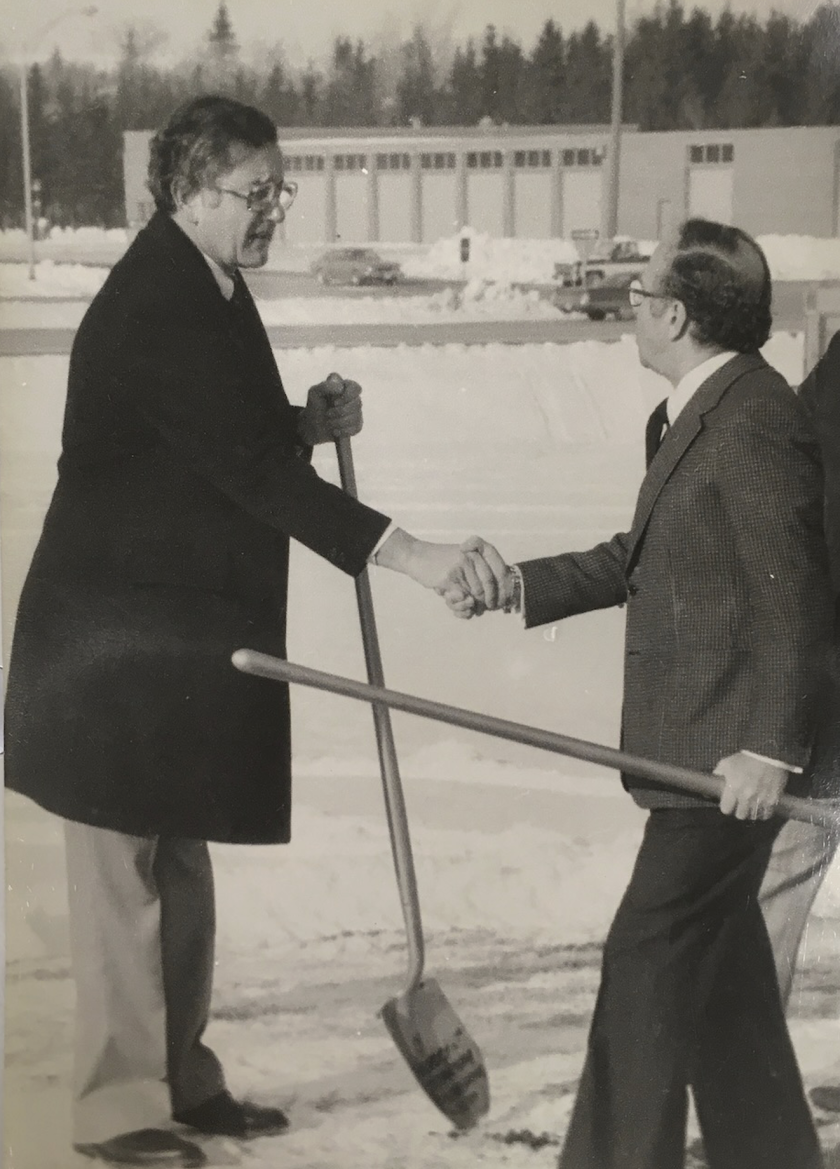 Two middle-aged white men, each holding a shovel, shake hands in a snowy landscape.