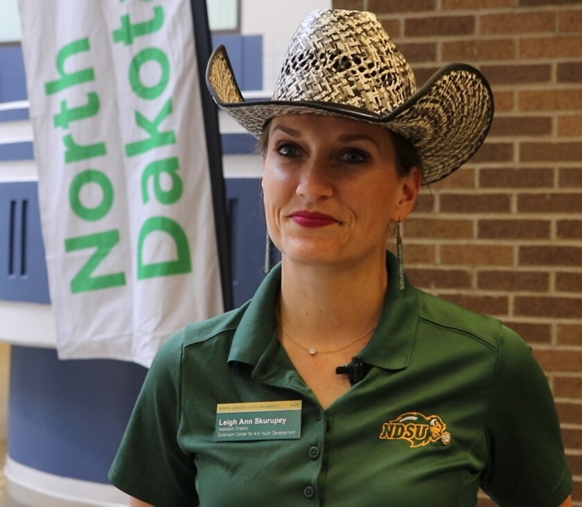 A woman wearing a green North Dakota State University polo shirt and a straw cowboy hat stands in front of a North Dakota 4-H sign