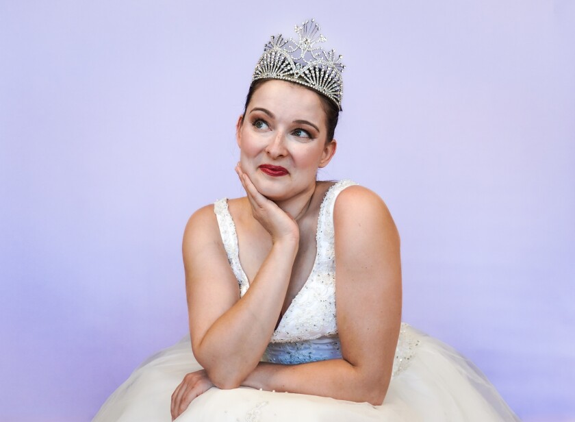 Light-skinned woman dressed in white princess outfit smiles and puts right hand to face, posed against light purple studio backdrop.
