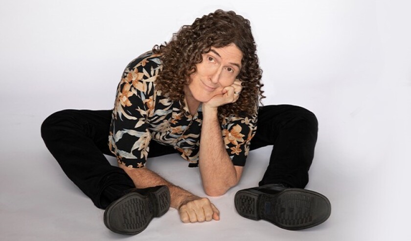 Studio portrait of "Weird Al" Yankovic sitting against a white backdrop, leaning forward over his spread legs with a whimsical expression on his face.