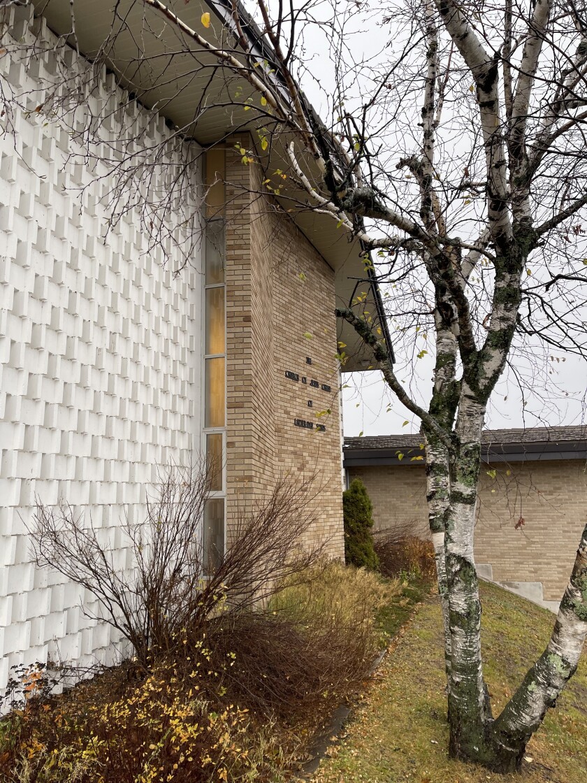 Exterior view of a church building on an overcast day, with ribbed concrete wall section at left and brick wall section at center. To the right is a tree with white bark and multiple branching trunks.