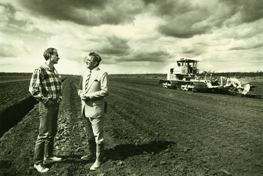 Two men stand in peat field, talking, at left. One is dressed in jeans and flannel, the other in a business suit with rubber boots. A peat extraction machine is visible in the background, to right.