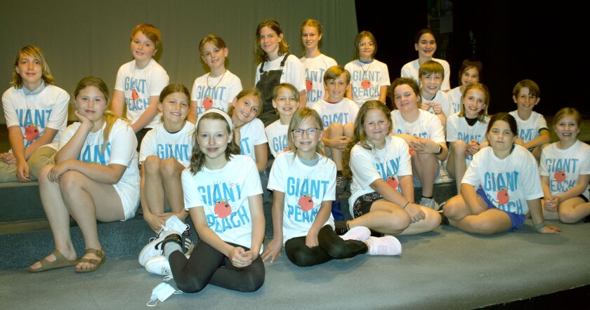 A large group of children sit together on risers, smiling and wearing t-shirts reading GIANT PEACH