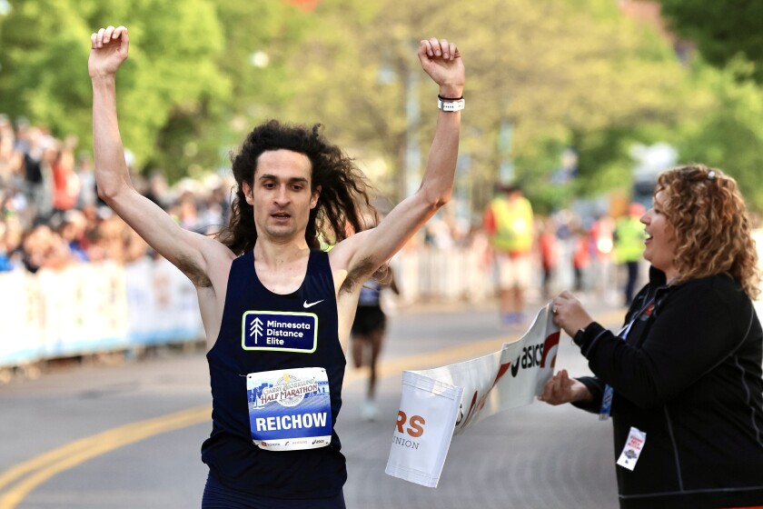 man with light skin and long brown hair raises arms over head at finish line