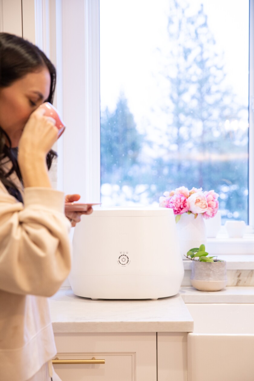 A woman sips coffee as she walks by a Lomi on the counter with a winter scene out the window. 