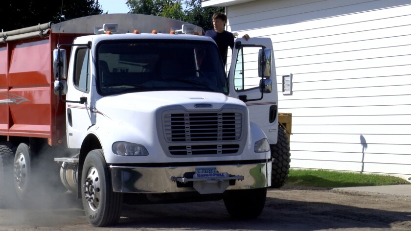 A man wearing a blue t-shirt stands on the platform of a white truck with a red box.