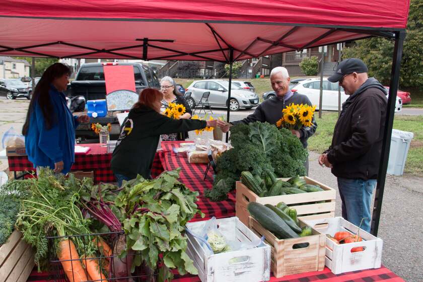 At a produce vendor stand, a woman takes payment from a man bearing an armload of sunflowers. In the foreground are boxes with carrots, cucumbers, and tomatoes among other fresh-grown foods.