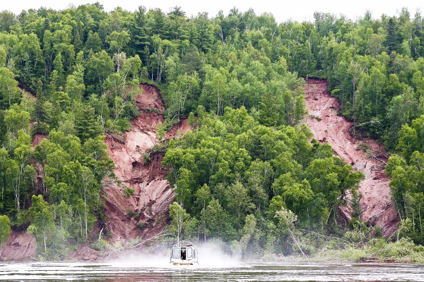 Three mudslides mark a hill otherwise covered in green trees, next to a river where an airboat is seen