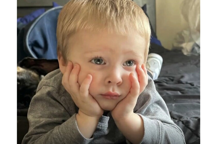A white toddler with blonde hair poses for a photo with his elbows resting on a bed and hands holding his face.