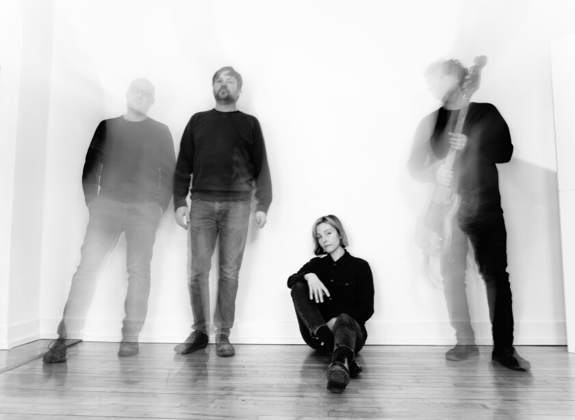 Black and white image of woman sitting amidst three standing men; the men are moving and blurred, while the woman looks directly into the camera.