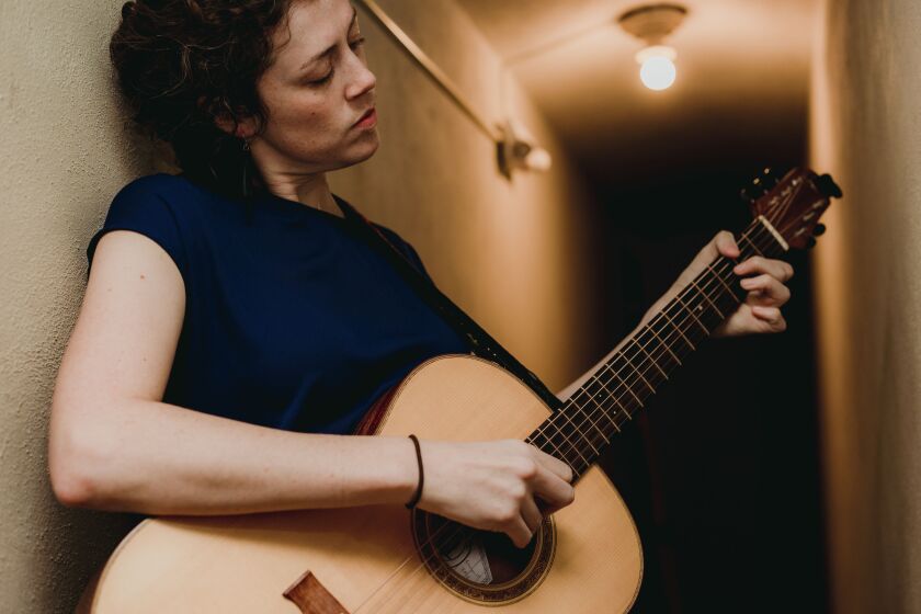 Rachel Kilgour thoughtfully plucks an acoustic guitar, in tight shot set in hallway with bare lightbulb visible in background.