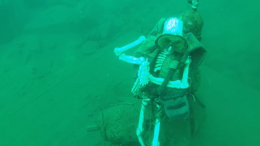 A skeleton dressed in scuba gear sits on a green lake bottom under water.