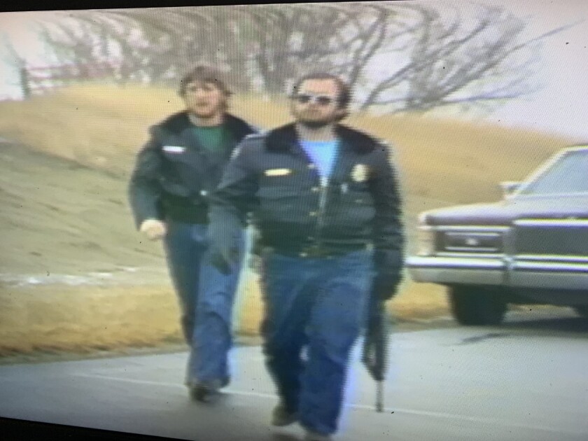 Two men in blue jeans and black police coats walk on the road. The man on the right wears dark aviators and carries a long gun.