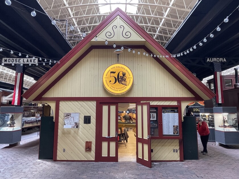 A wooden building painted yellow with burgandy trim stands in an enclosed train shed. An illuminated "50" golden seal is mounted on the exterior.