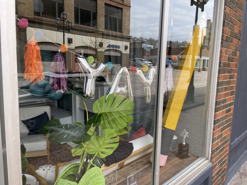 A storefront window features silver balloons reading "YAY" hanging from a string, next to an outsized model pencil.