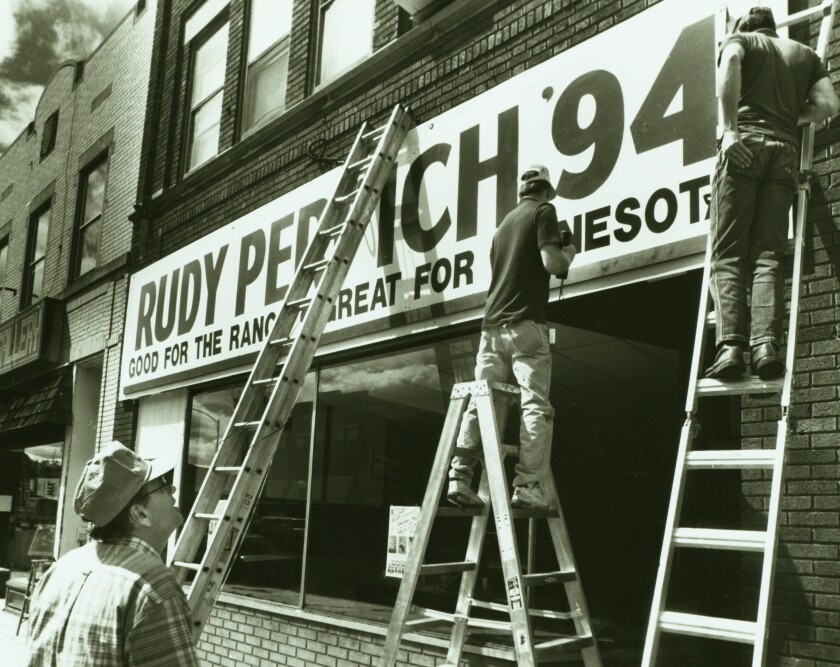 Three men with ladders apply a sign to a storefront. The sign reads, "RUDY PERPICH '94: GOOD FOR THE RANGE, GREAT FOR MINNESOTA."