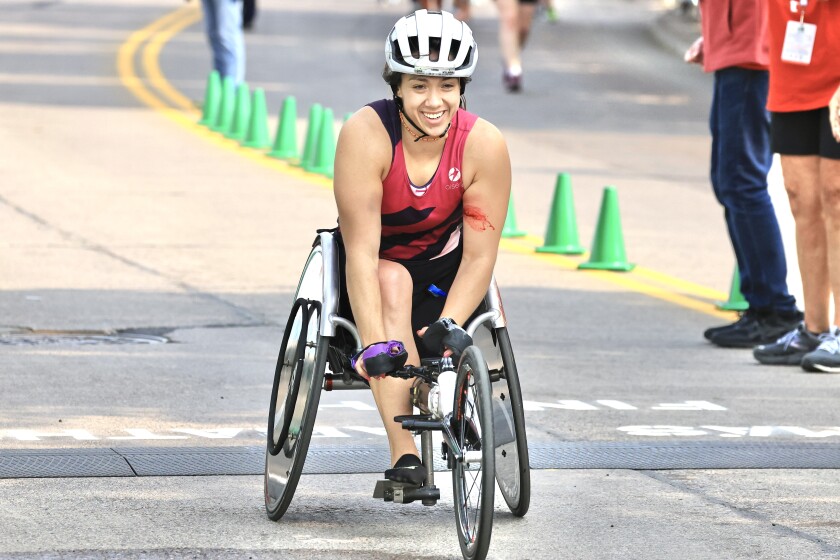 woman in helmet and red and black top rides three-wheeled wheelchair past finish line