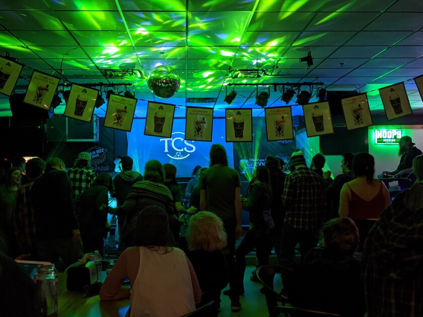 A crowd gathers in front of a green-lit stage in a barroom. Due to the crowd, the stage itself is not visible.