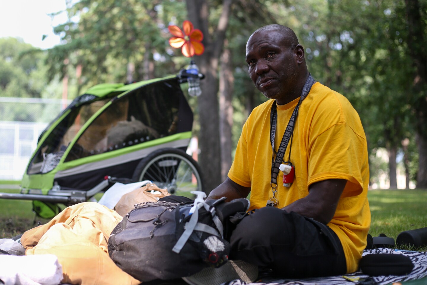 A man in a yellow T-shirt sits on a blanket surrounded by bags with a bike trailer behind him in a shady park. He wears a lanyard with an inhaler on it around his neck.
