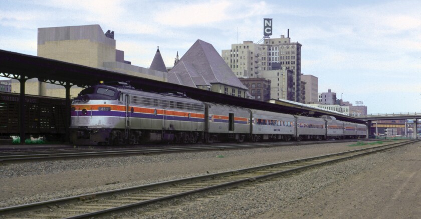 An silver Amtrak train with red-and-blue lines sits outside a 19th-century train depot. A city skyline rises in the background, with empty tracks in the foreground.