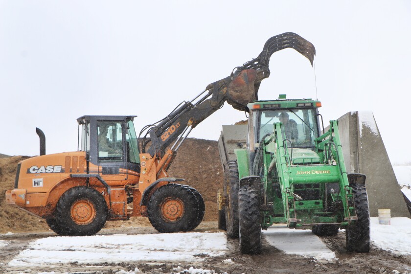 A loader (left) puts silage and feed components into a trailer, pulled by a John Deere green and yellow tractor at right.
