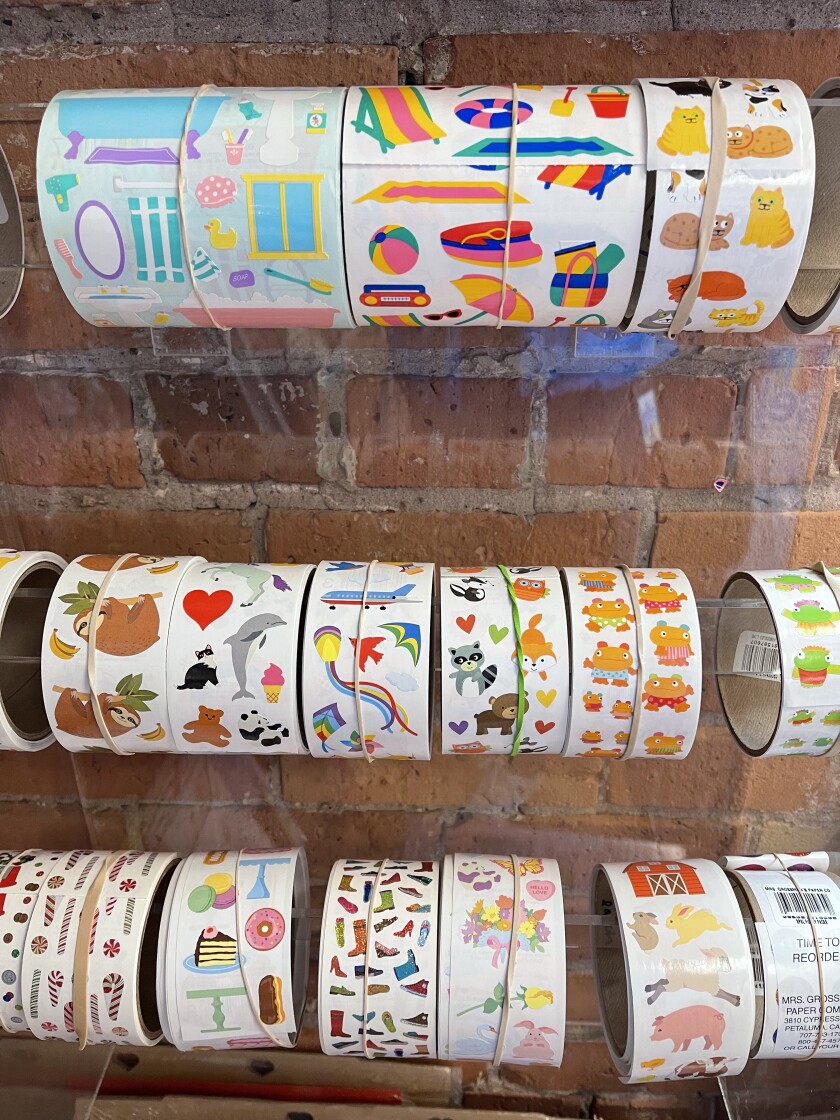 Rows of sticker rolls — featuring imagery like cake, farm animals, and zoo animals — against a brick wall in a retail shop.