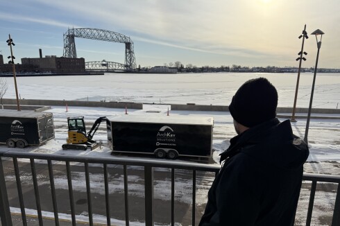 Man in black jacket and winter hat is seen from behind, looking over lake harbor with lift bridge in background. Railing stands behind him.