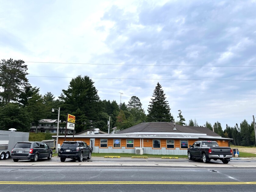 Exterior view of a low, octagonal restaurant with log sides. A boreal forest is visible in the background, and a few large black vehicles are parked outside.