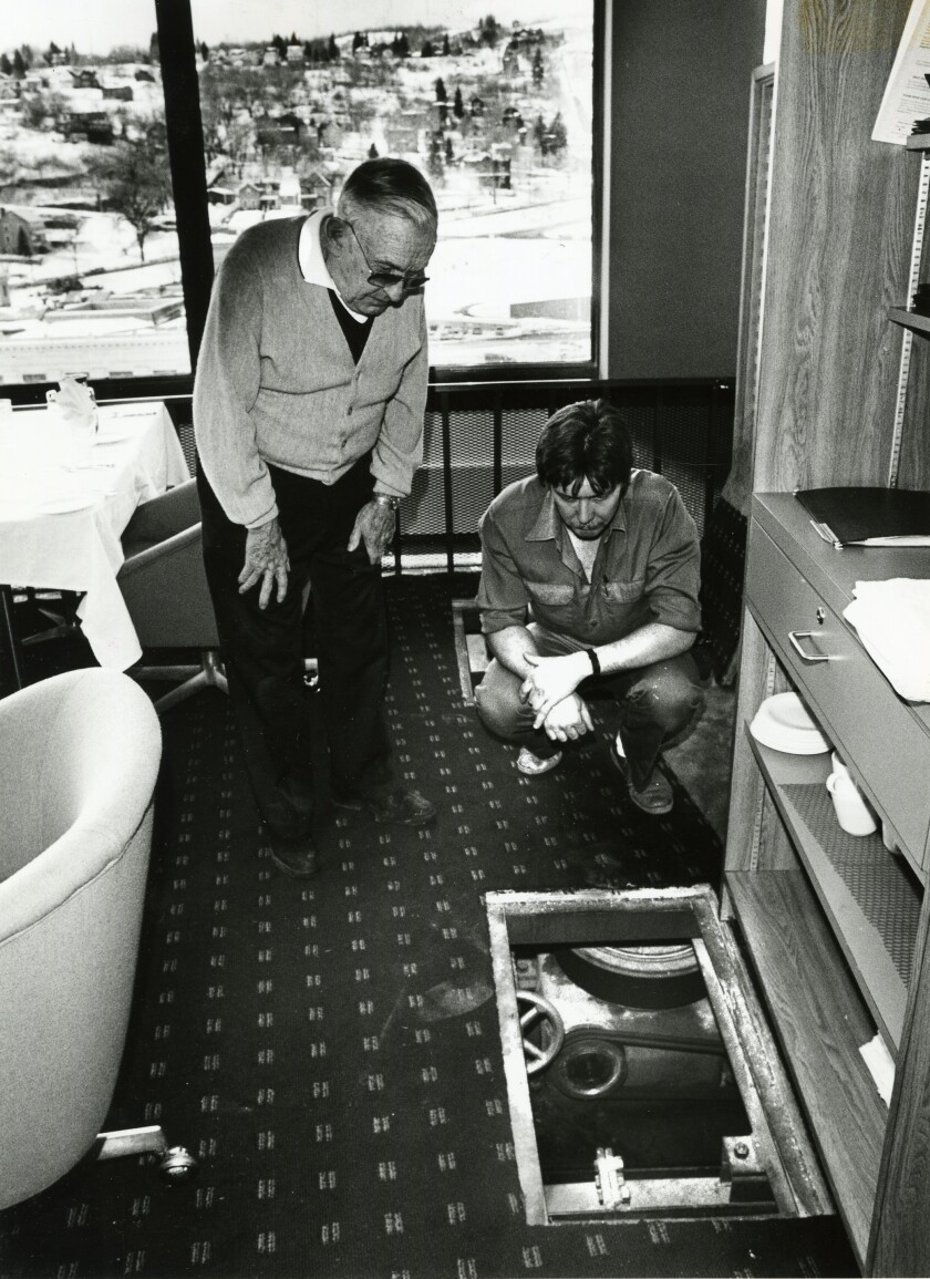 Black and white image of two men crouched over a hole in a restaurant floor. Through the hole, a pulley mechanism can be seen.