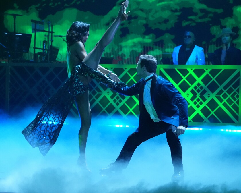 A woman in long green gown swings her leg high above the head of a crouching man while dancing on a green-lit, vapor-filled stage.
