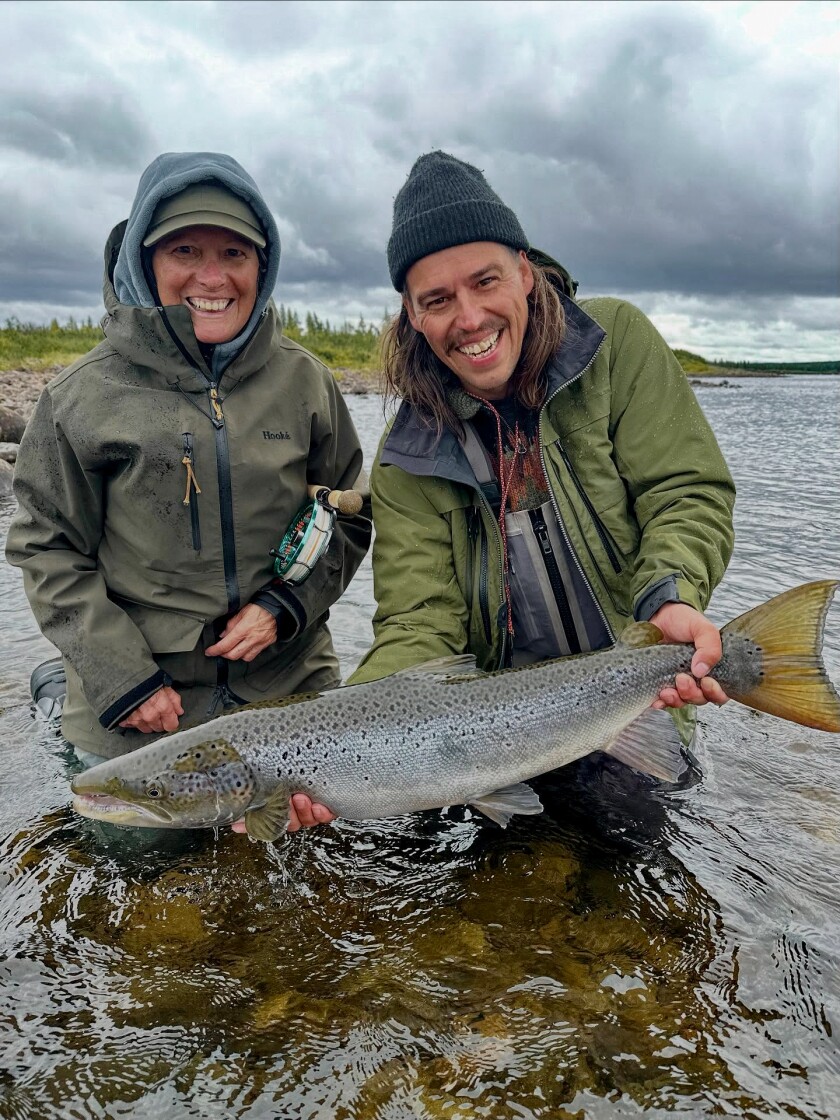 Two light-skinned people dressed from head to toe in green fishing gear stand thigh-deep in a stream under grey clouds, holding a large fish and smiling.