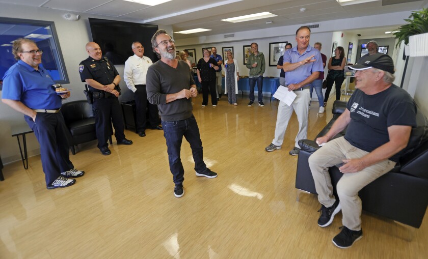 A bearded man and a man in a black T-shirt and baseball cap laugh while a third man gestures.