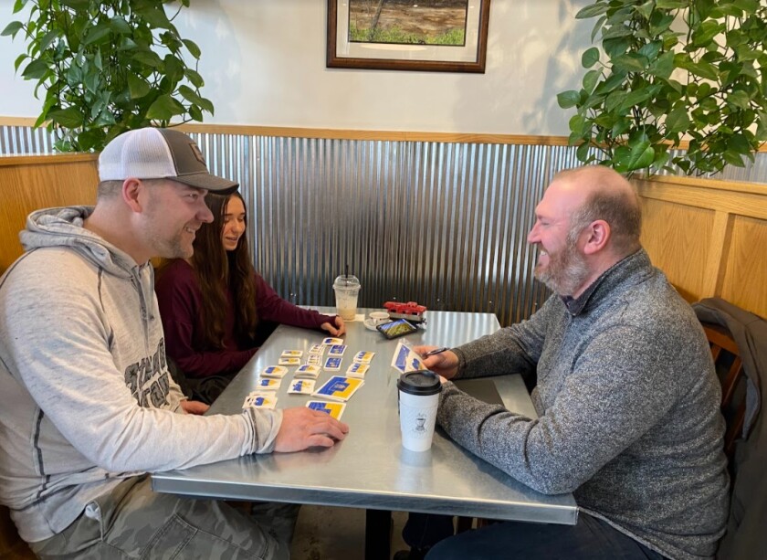Anastasiia Biermaier, her uncle Dan Lundberg, and Seth Neistadt sit in a booth with stickers on the table.