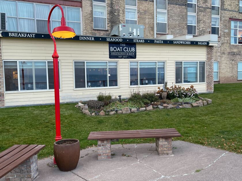 Exterior view of restaurant bearing sign "The Boat Club." In the foreground is a red streetlamp and a brown bench, with landscaping in the middle ground.