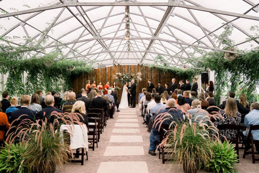 Plants hang from metal rails on the ceiling above guests seated during a wedding ceremony. At the altar is a backdrop of vertical weathered wood boards in various shades of brown.
