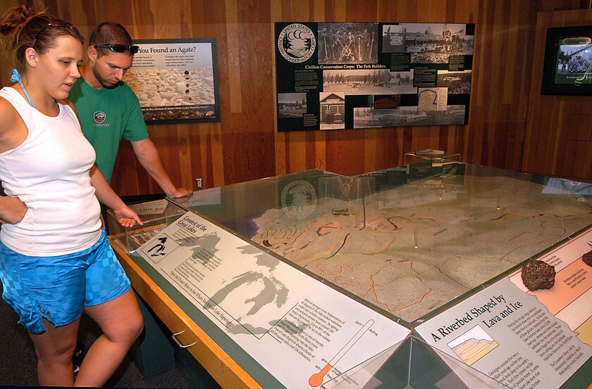 A light-skinned woman and light-skinned man, both in casual warm weather clothing, peruse a map display in a room with natural wood walls.
