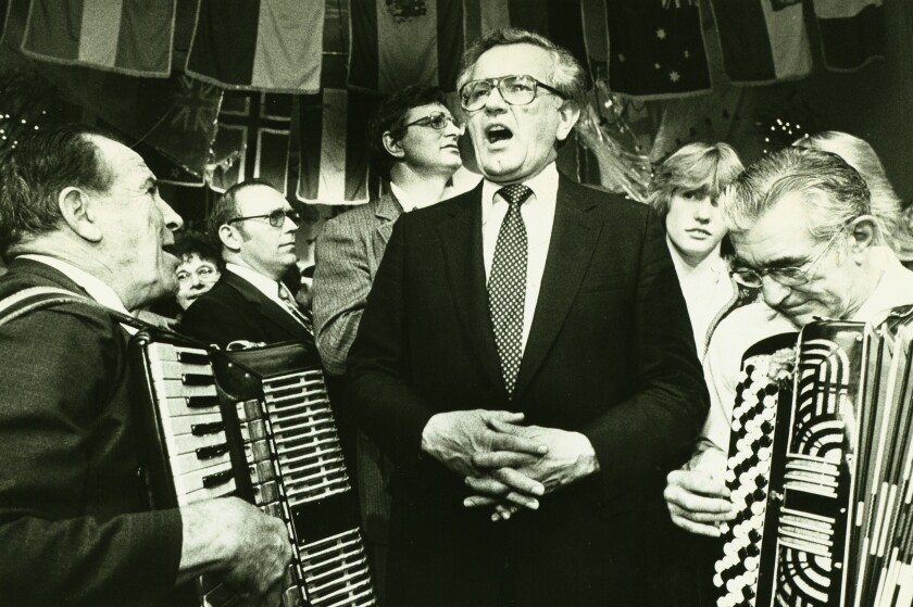 Man in business suit sings emphatically as two older men playing accordions stand astride him.