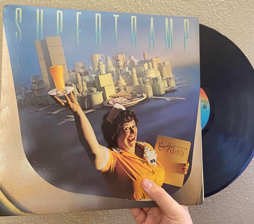 A hand holds a copy of Supertramp's "Breakfast in America" on vinyl, with cover image of diner server in Statue of Liberty pose.