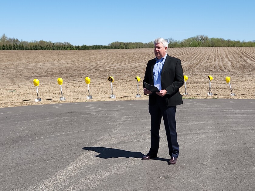 A man in a suit stands in front of a dirt covered field and shovels topped with yellow hard hats.