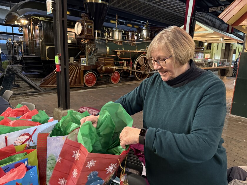 Light-skinned woman of senior age smiles as she holds Christmas gift bags stuffed with red and green tissue paper, with steam locomotive visible in background.