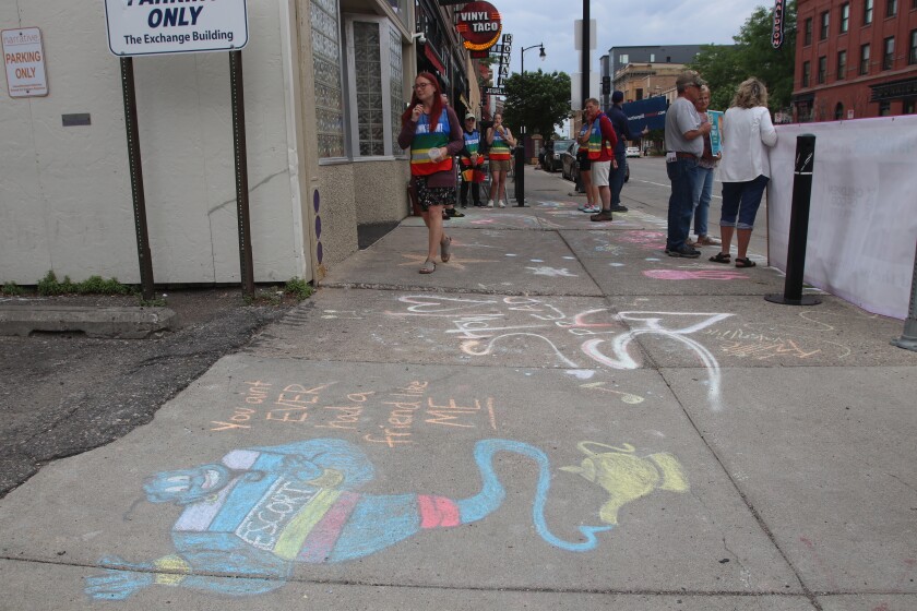 A woman in a rainbow vest walks in front of abortion clinic, with protesters to her left and a colorful chalk drawing of a genie in a bottle on the sidewalk