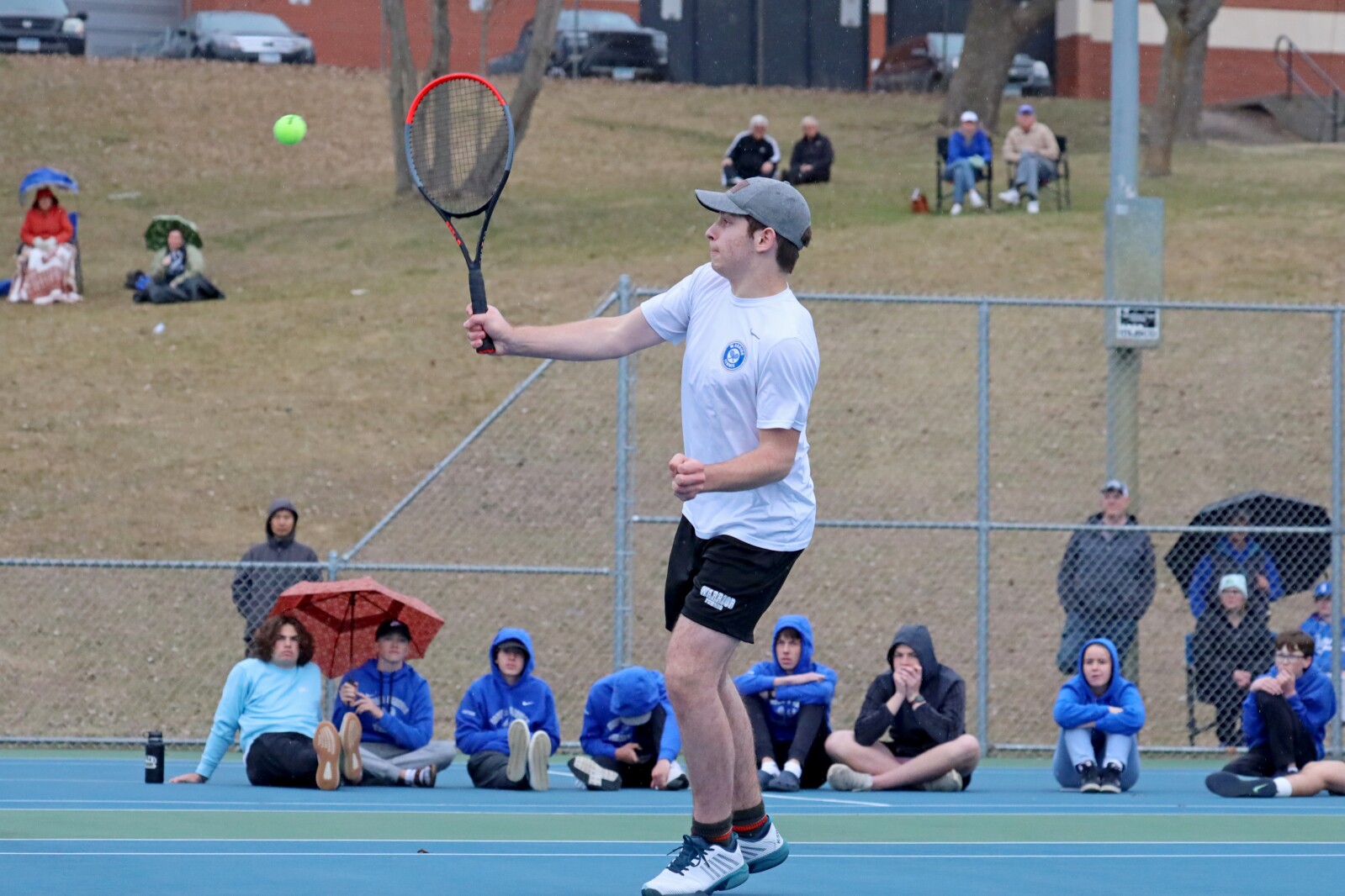 Tennis player hits the ball on the court. Fans watch in the background.