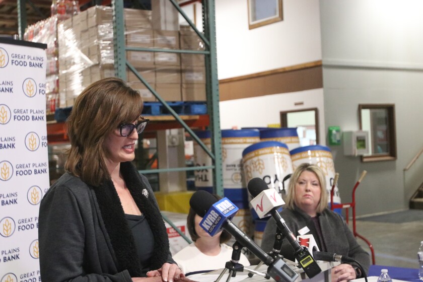 A woman in a dark gray outfit and large glasses speaks at a podium with media microphones on it.