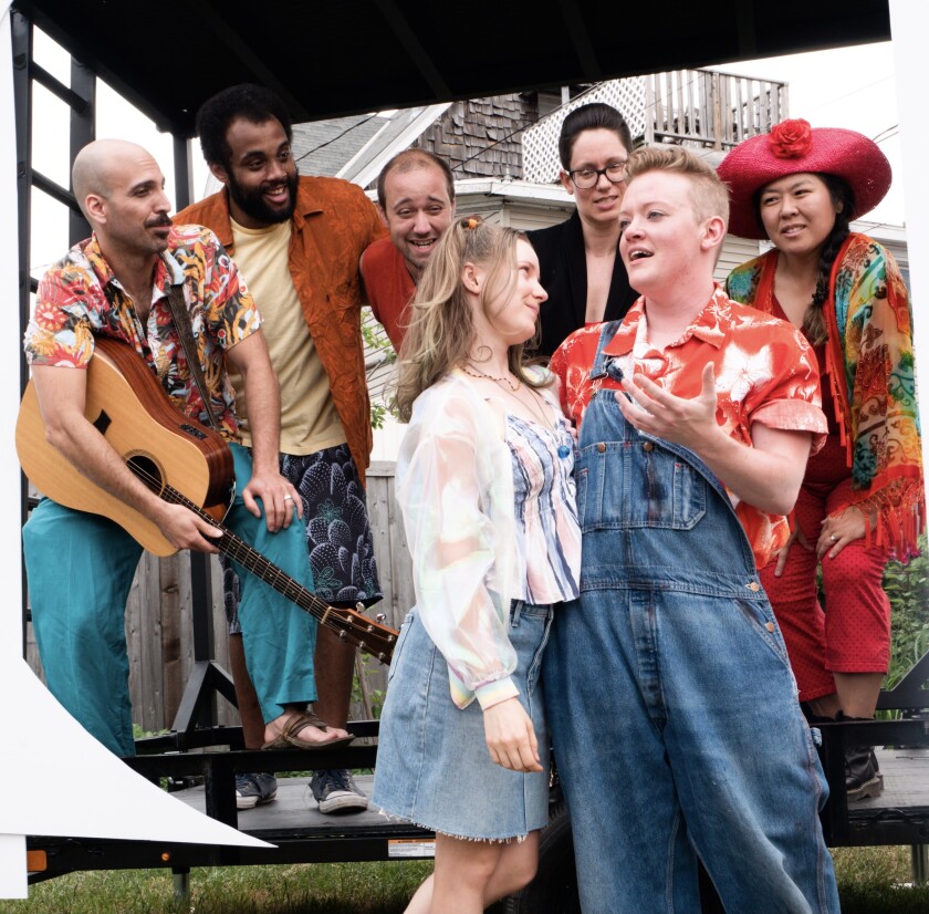 A colorfully dressed ensemble of actors look on as two people stand in the middle, arm in arm, sharing a tender moment.