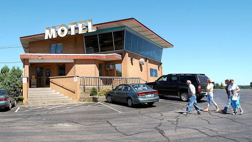 A family of people walk toward a brick building topped with a glass-walled space under a roof with a large sign reading, "MOTEL."