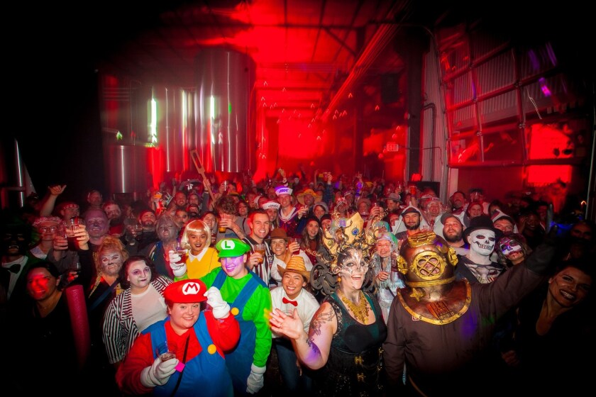 Large crowd of costumed people in red-lit brewery, with large fermentation tanks visible in background.