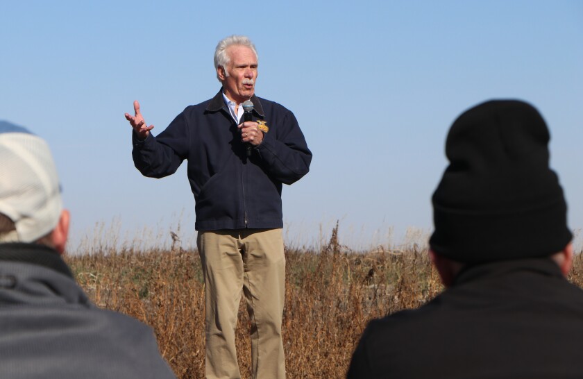 A white-haired man speaks from an outdoor platform as listeners are in silhouette in the foreground.