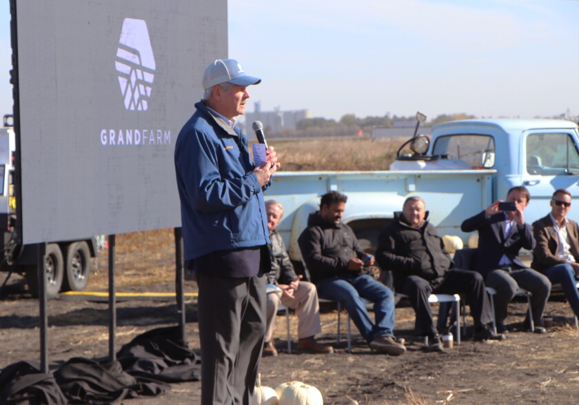 A man speaks in a blue Grand Farm ball cap, flanked by dignitaries.